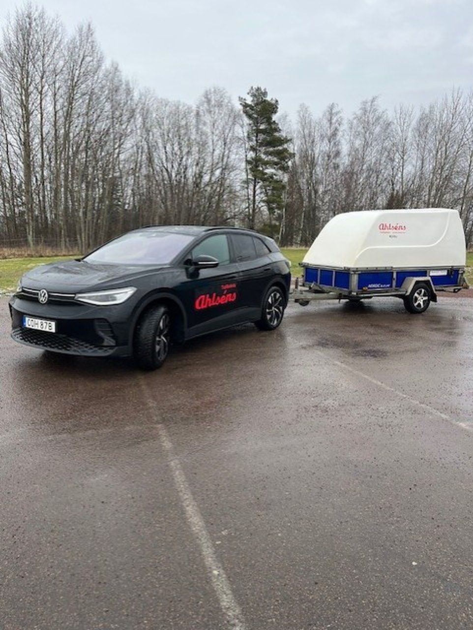 Svart VW ID.4 med Ahlséns dekor och ett kåpsläp på en stor parkering