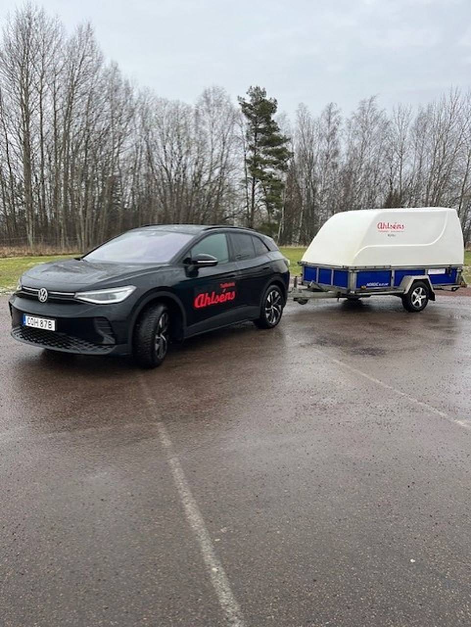 Svart VW ID.4 med Ahlséns dekor och ett kåpsläp på en stor parkering