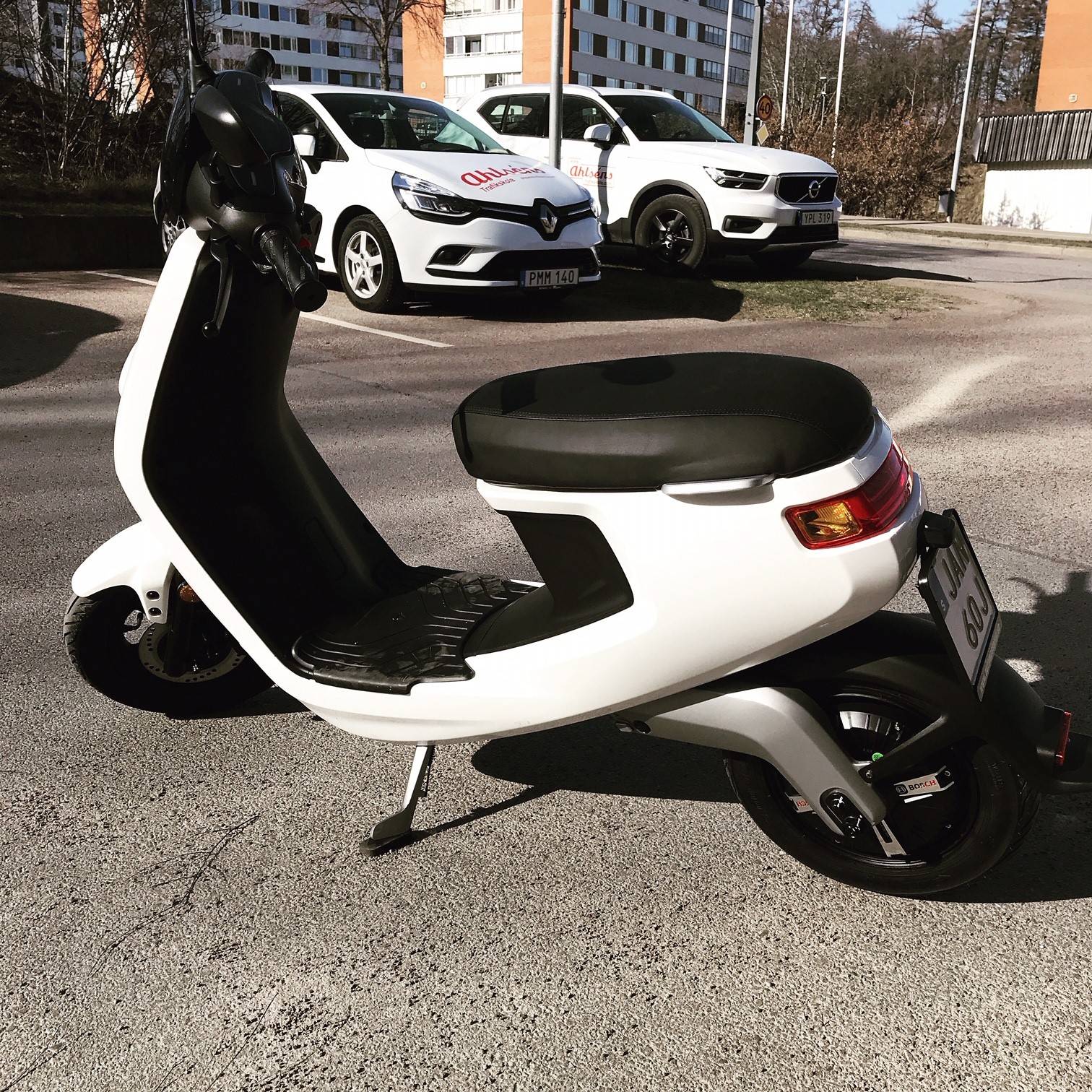Vit elmoped på en parkering. I bakgrunden står två vita övningsbilar med Ahlséns dekor