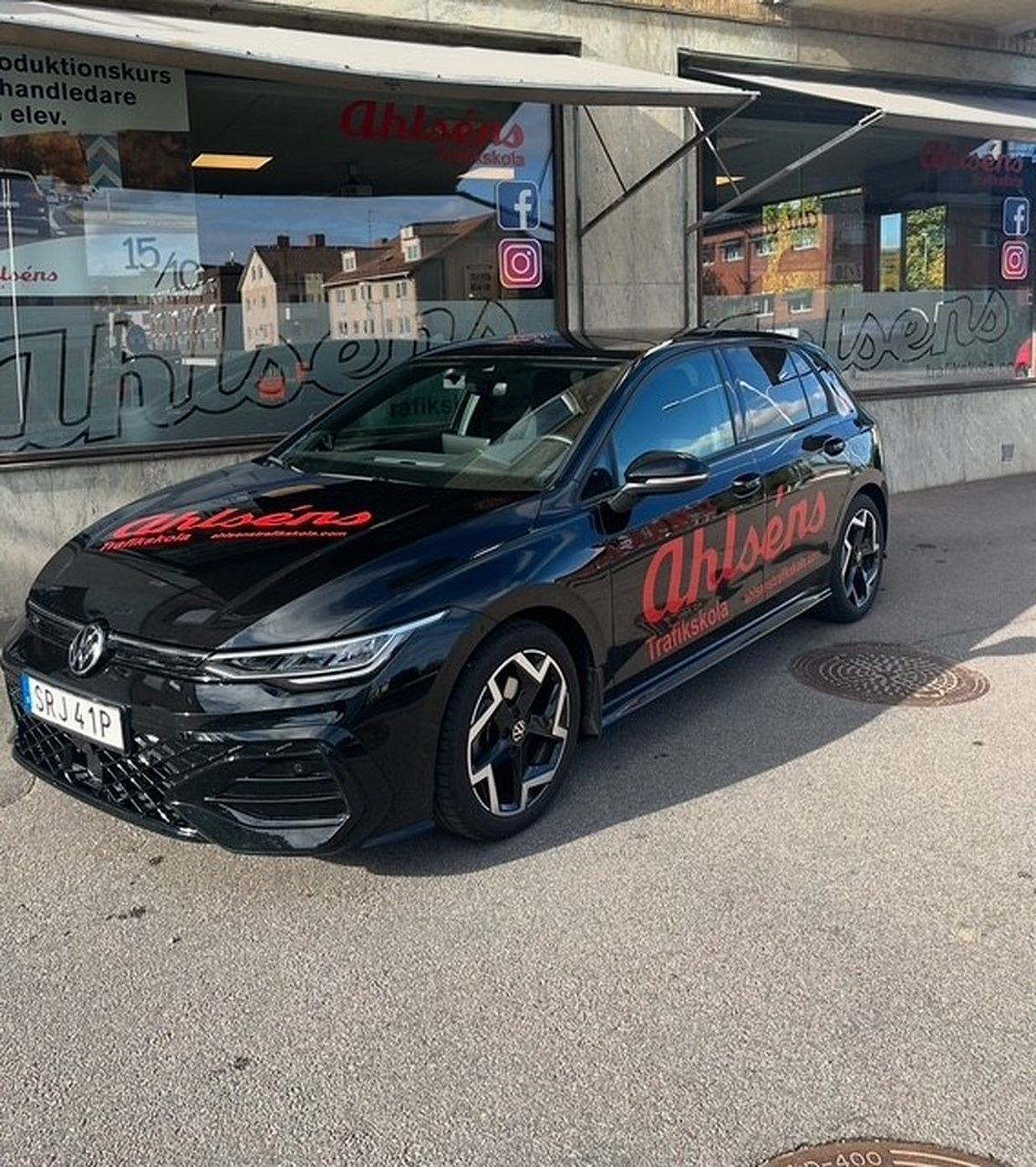 Svart VW Golf med Ahlséns röda dekor parkerad framför trafikskolans skyltfönster i Mjölby