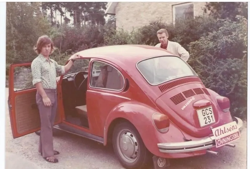 Ett gammalt färgfoto av en röd VW Bubbla med skylten Ahlséns övningsbil. En ung man står vid den öppna dörren och en man skymtar bakom bilen.